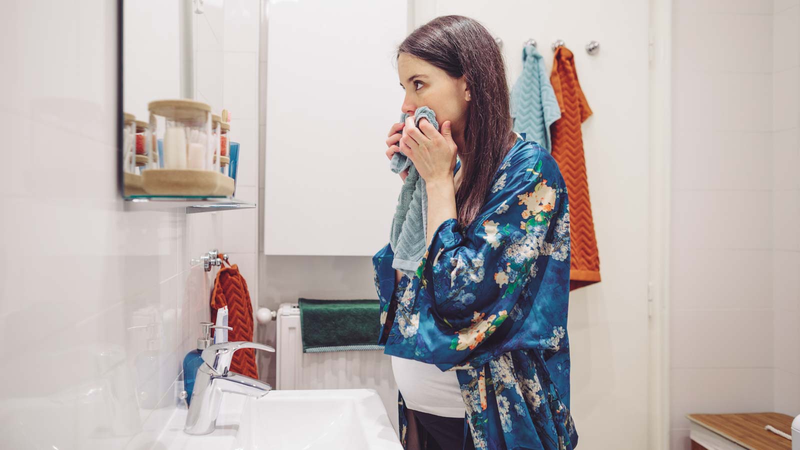 nauseous pregnant woman standing at a bathroom sink in her home holding a towel to her mouth